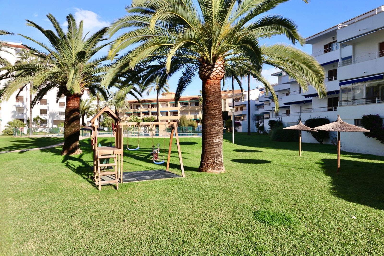Lush communal garden featuring a children's playground with swings, mature palm trees, and two straw parasols, set against residential buildings under a clear blue sky.
