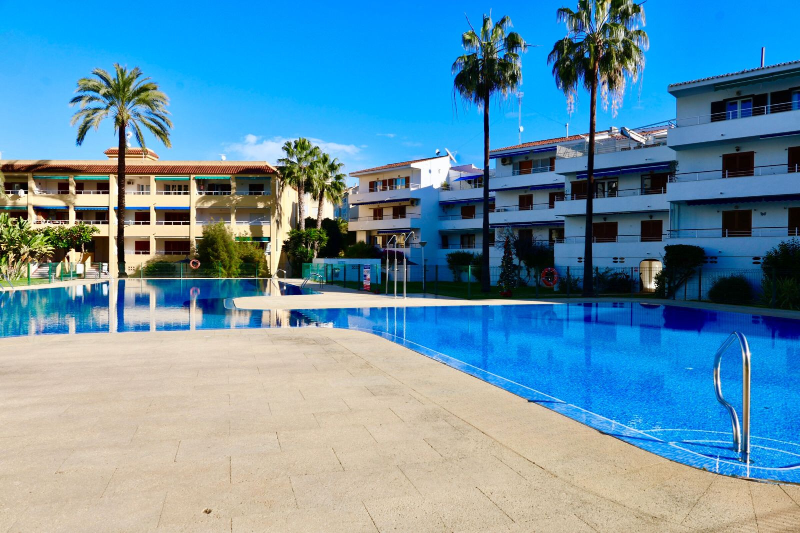 Spacious communal swimming pool with blue mosaic tiles, bordered by a light-tiled patio and palm trees. Apartment buildings with balconies and blue awnings are visible under a clear sky.
