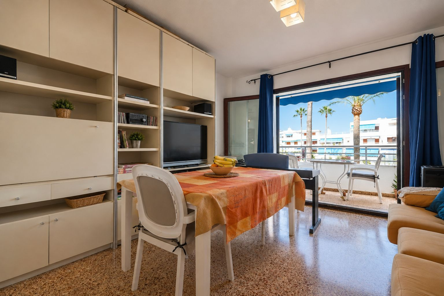 The bright living-dining area features a built-in media unit and a dining table, opening directly onto a terrace with palm tree views and distant white buildings.