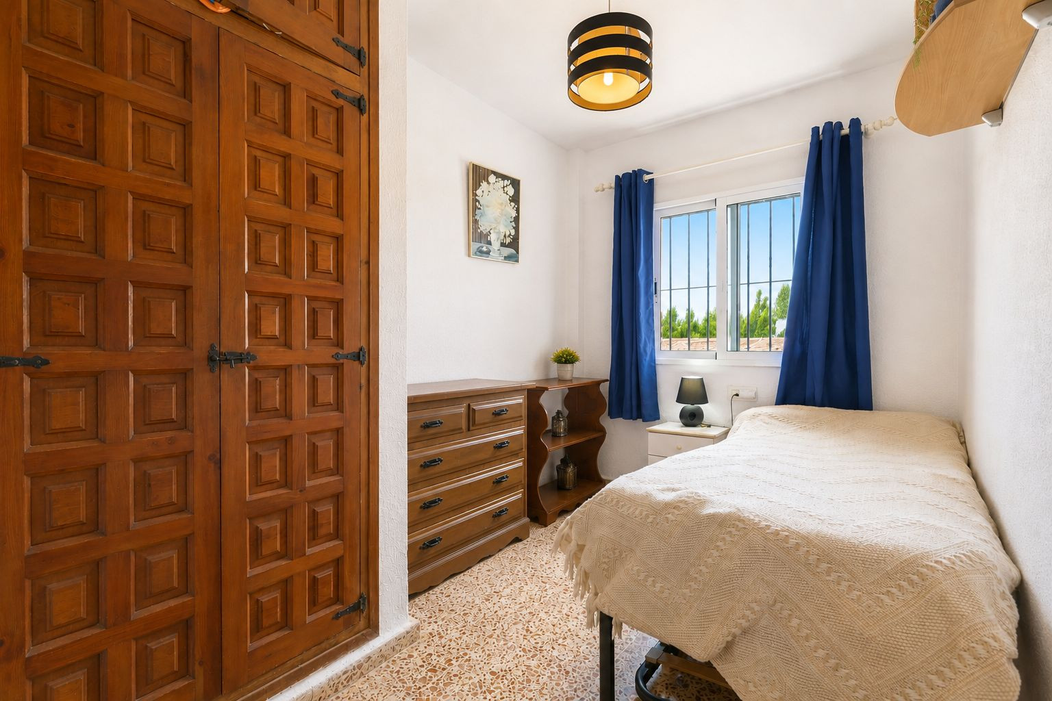 This cozy bedroom features a prominent built-in wooden wardrobe with decorative paneling and traditional black hardware. Natural light enters through a window with blue curtains, offering views of surrounding greenery.