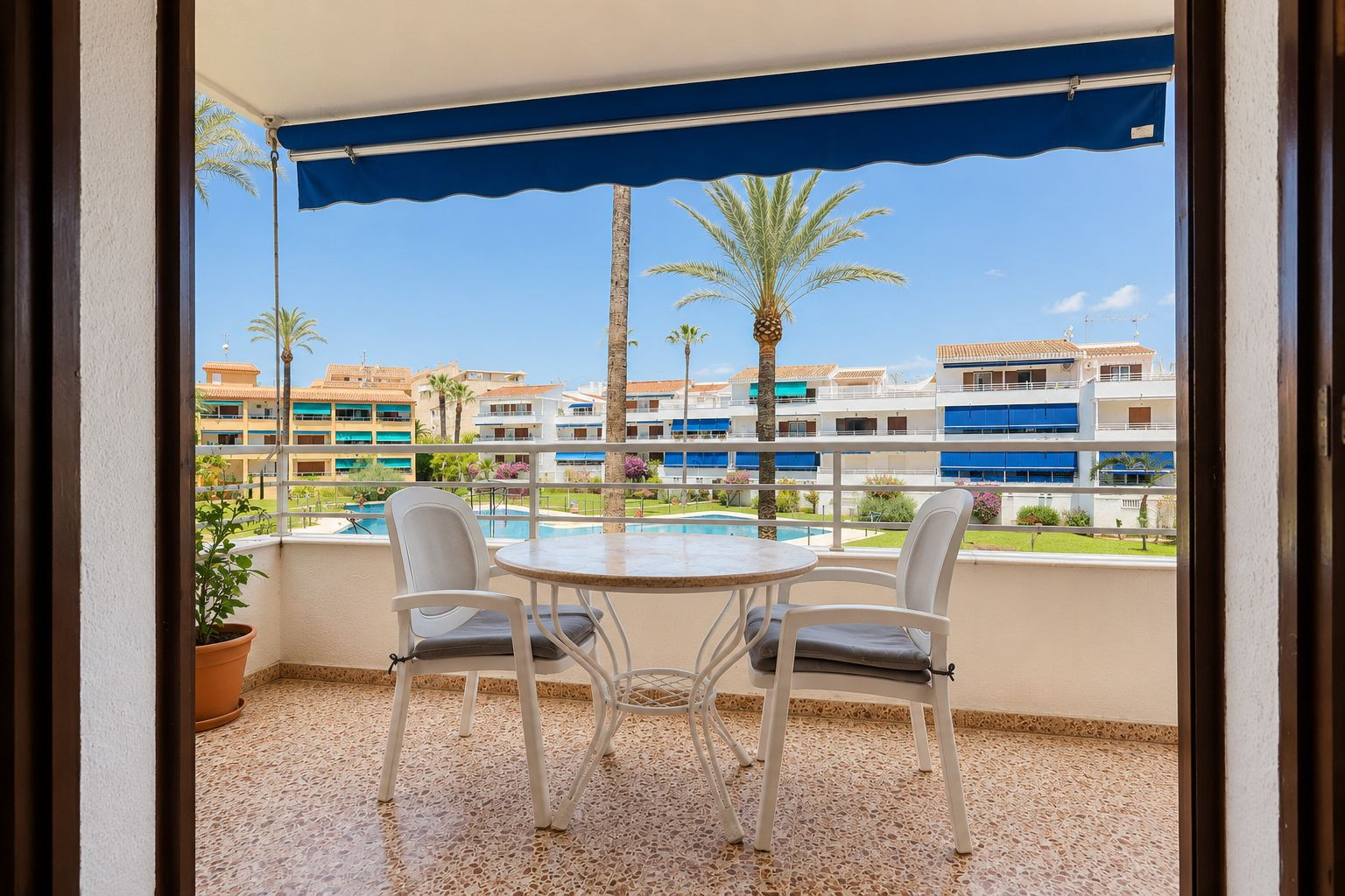 Tiled terrace featuring a round table and two chairs, shaded by a blue awning. It overlooks the communal pool, landscaped gardens, and palm trees.