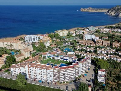 Aerial view of a coastal urbanization featuring multiple residential buildings, communal pools, and green areas, adjacent to a sandy beach and the Mediterranean Sea, with a distant rocky cape.