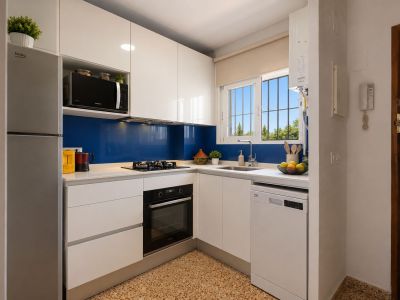 Modern and functional kitchen featuring glossy white cabinetry, a light countertop, and a vibrant blue backsplash. Equipped with integrated appliances and a window overlooking greenery.