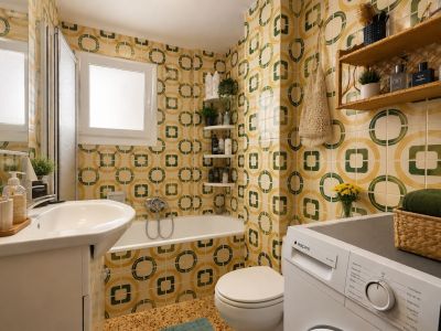 The full bathroom features distinctive yellow and green geometric patterned tiles, a white vanity with sink, a bathtub with shower screen, and an integrated washing machine, illuminated by a frosted window.