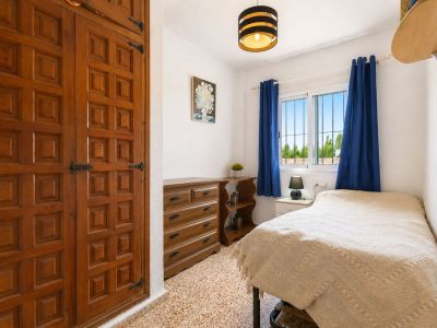 This cozy bedroom features a prominent built-in wooden wardrobe with decorative paneling and traditional black hardware. Natural light enters through a window with blue curtains, offering views of surrounding greenery.