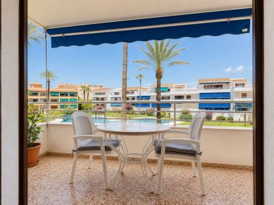 Tiled terrace featuring a round table and two chairs, shaded by a blue awning. It overlooks the communal pool, landscaped gardens, and palm trees.