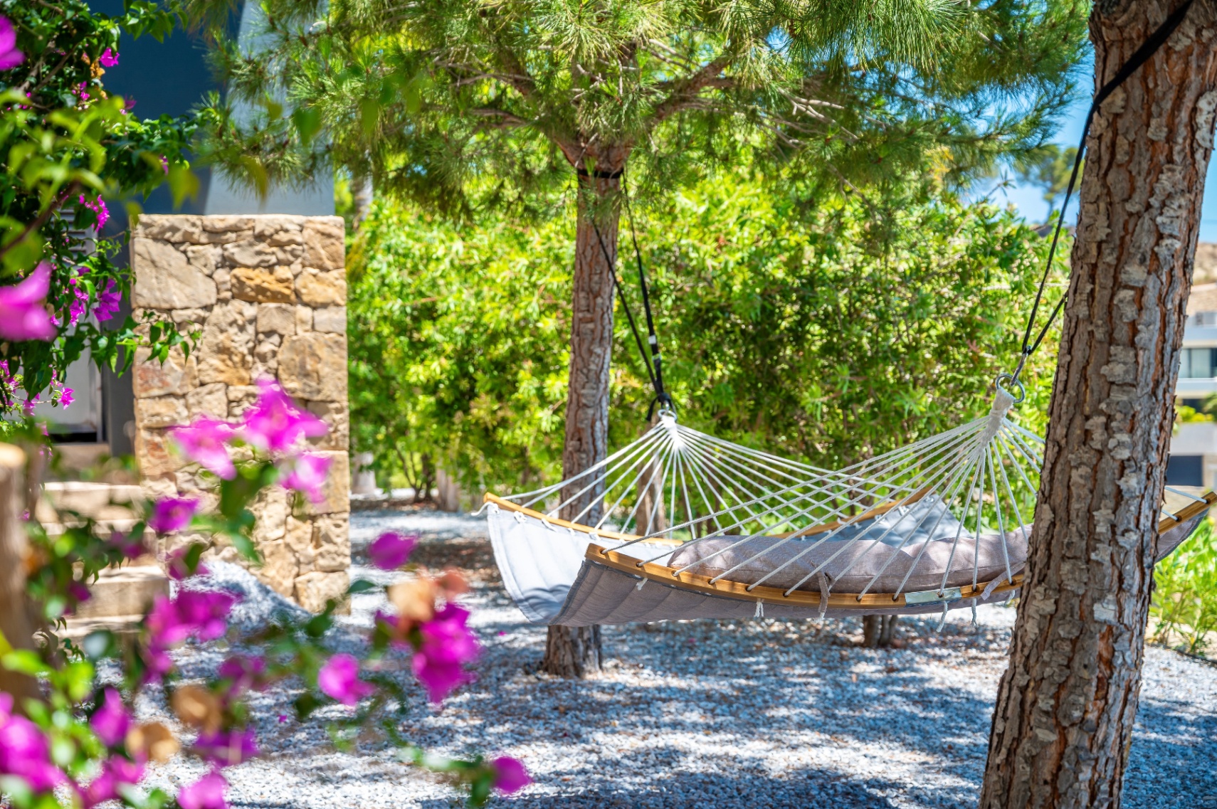 A tranquil garden corner featuring a comfortable grey hammock with wooden spreader bars, suspended between trees on a gravel bed, framed by vibrant bougainvillea.