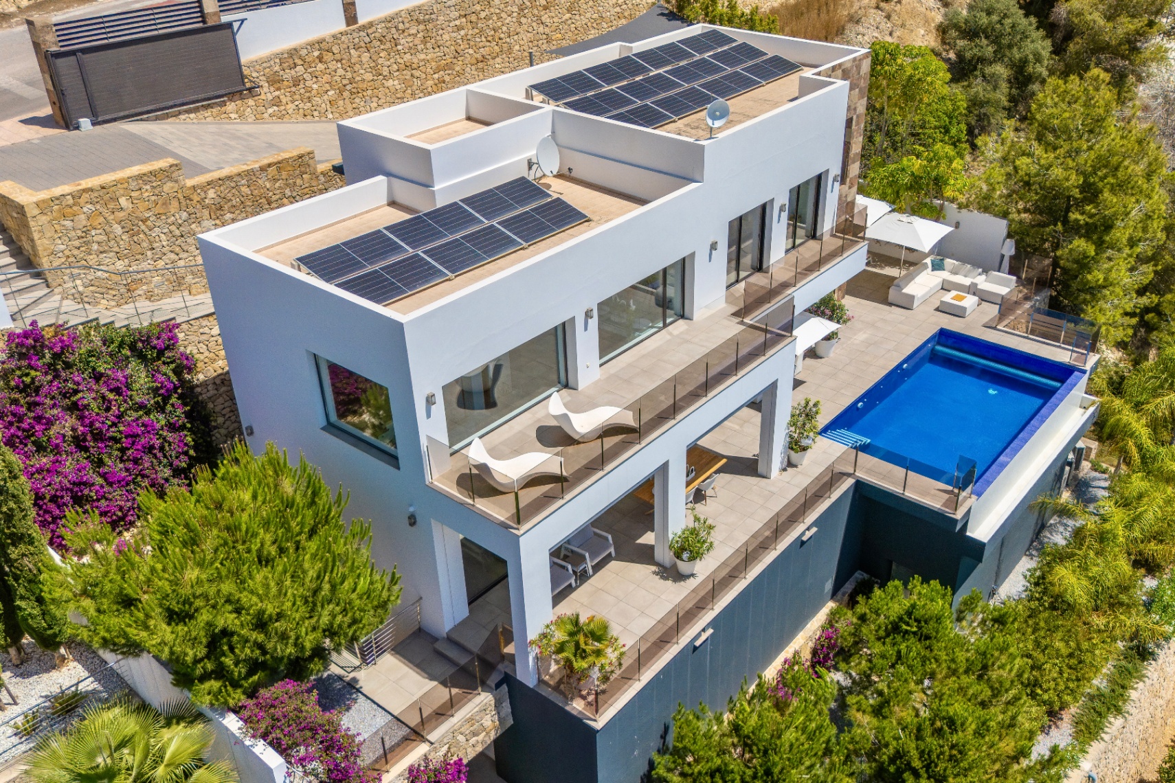 Aerial view of a modern white villa featuring multiple terraced levels, a rectangular swimming pool, and extensive outdoor lounge areas. Solar panels are visible on the flat roofs, set amidst lush Mediterranean landscaping.