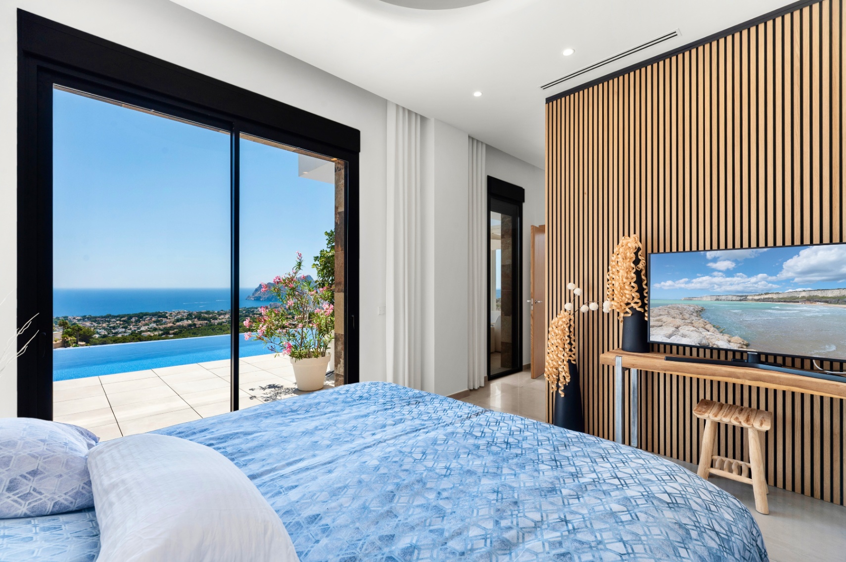 Bedroom featuring expansive sliding glass doors that open to an infinity pool and panoramic sea views. A modern slatted wood wall serves as a backdrop for the TV, complementing the light interior.