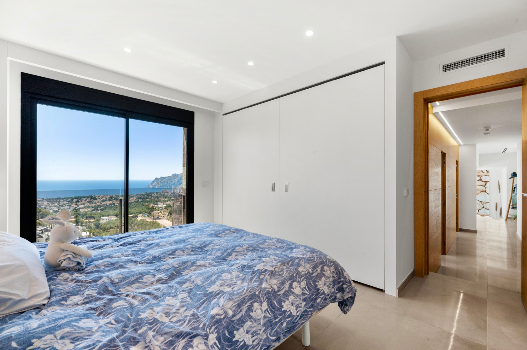 Bedroom with full-height sliding glass doors showcasing panoramic sea and town views. Features include integrated white wardrobes, recessed lighting, and a light tiled floor.
