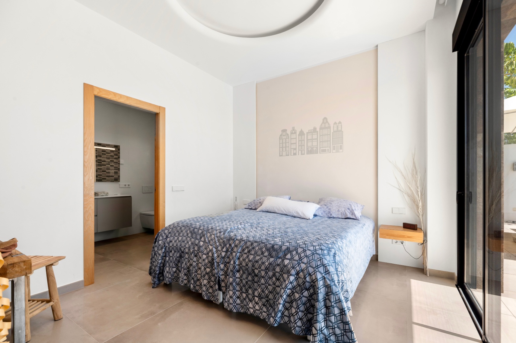 Bright bedroom featuring a double bed, an accent wall with building silhouettes, and a wooden-framed doorway to an en-suite bathroom. Direct access to a terrace via a large sliding glass door.