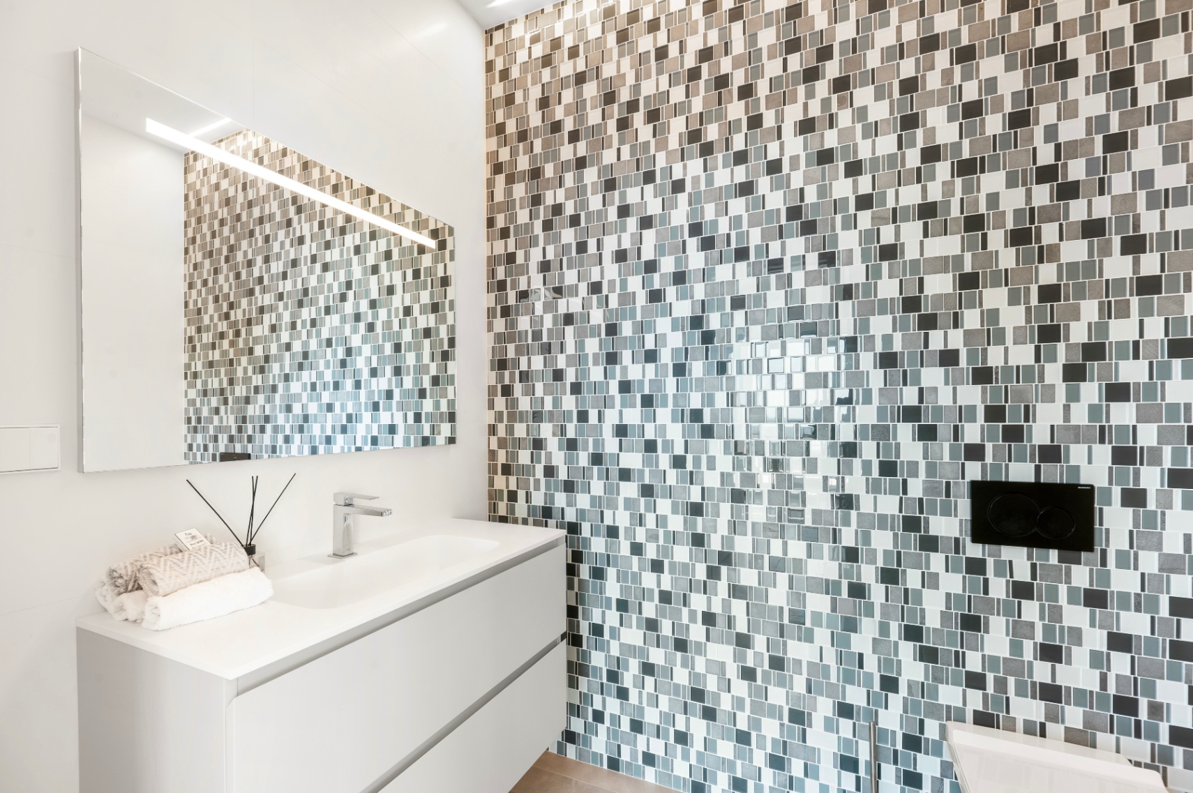 Contemporary bathroom featuring a white floating vanity with integrated sink and chrome faucet. The accent wall is clad in a grey and white mosaic tile, complemented by an illuminated mirror.