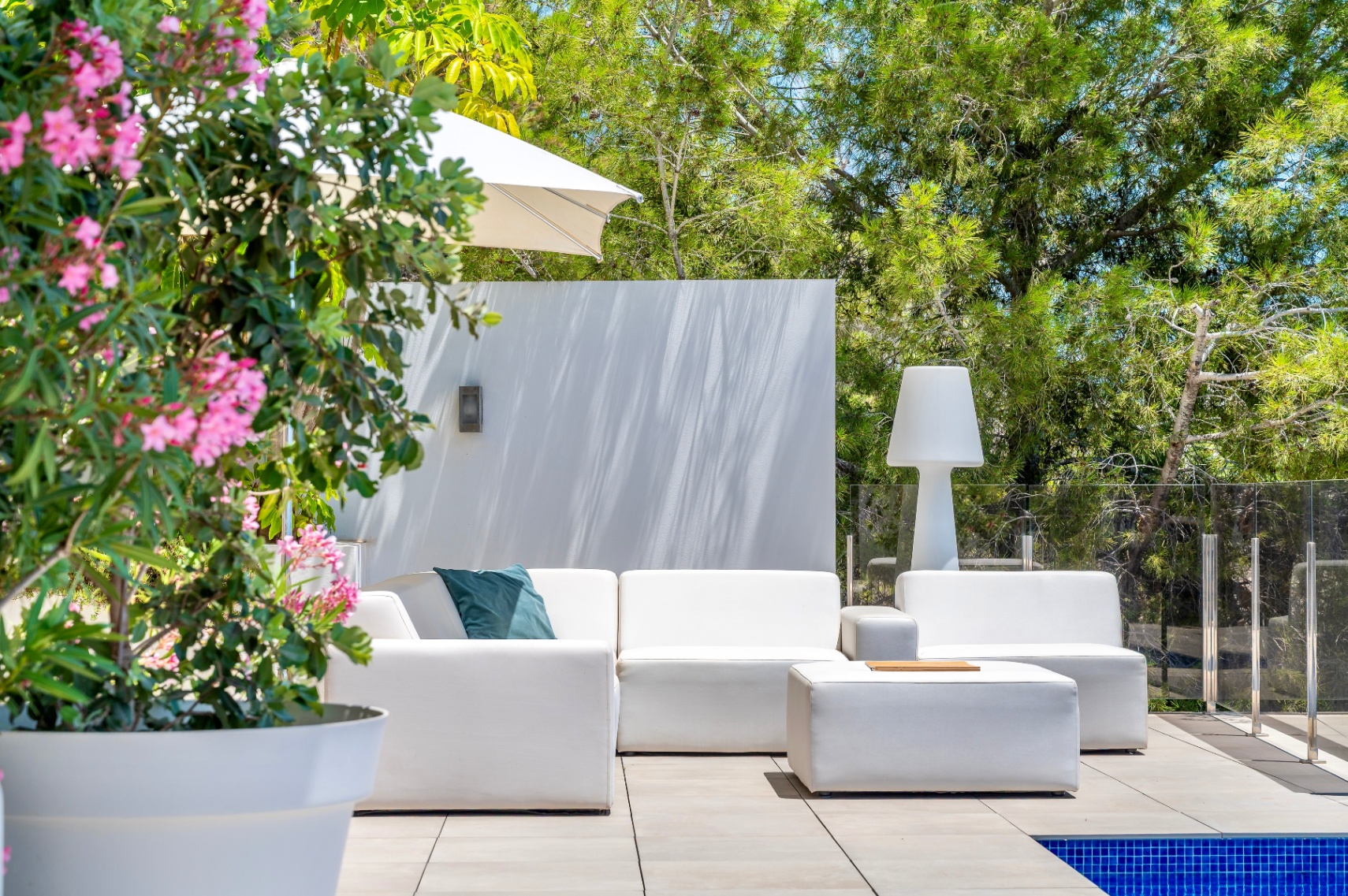Contemporary outdoor lounge area with white modular sofas and a matching ottoman on a tiled patio, bordered by a glass railing and lush greenery. A pink flowering plant adds a touch of color.