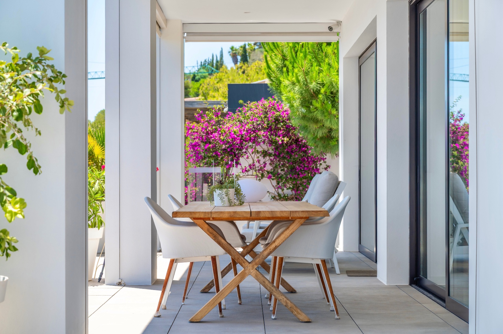 Covered outdoor dining terrace with a wooden table and white chairs, framed by lush greenery and vibrant bougainvillea, perfect for al fresco meals.