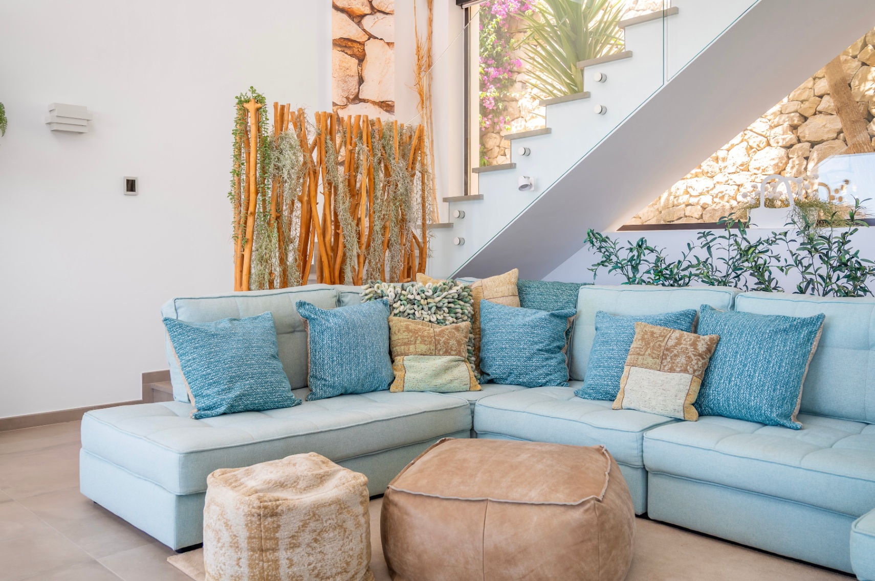 Cozy living area featuring a light blue sectional sofa, assorted cushions, and two leather poufs. A modern glass-railed staircase ascends alongside a natural wood and plant screen, with a stone wall visible beyond.