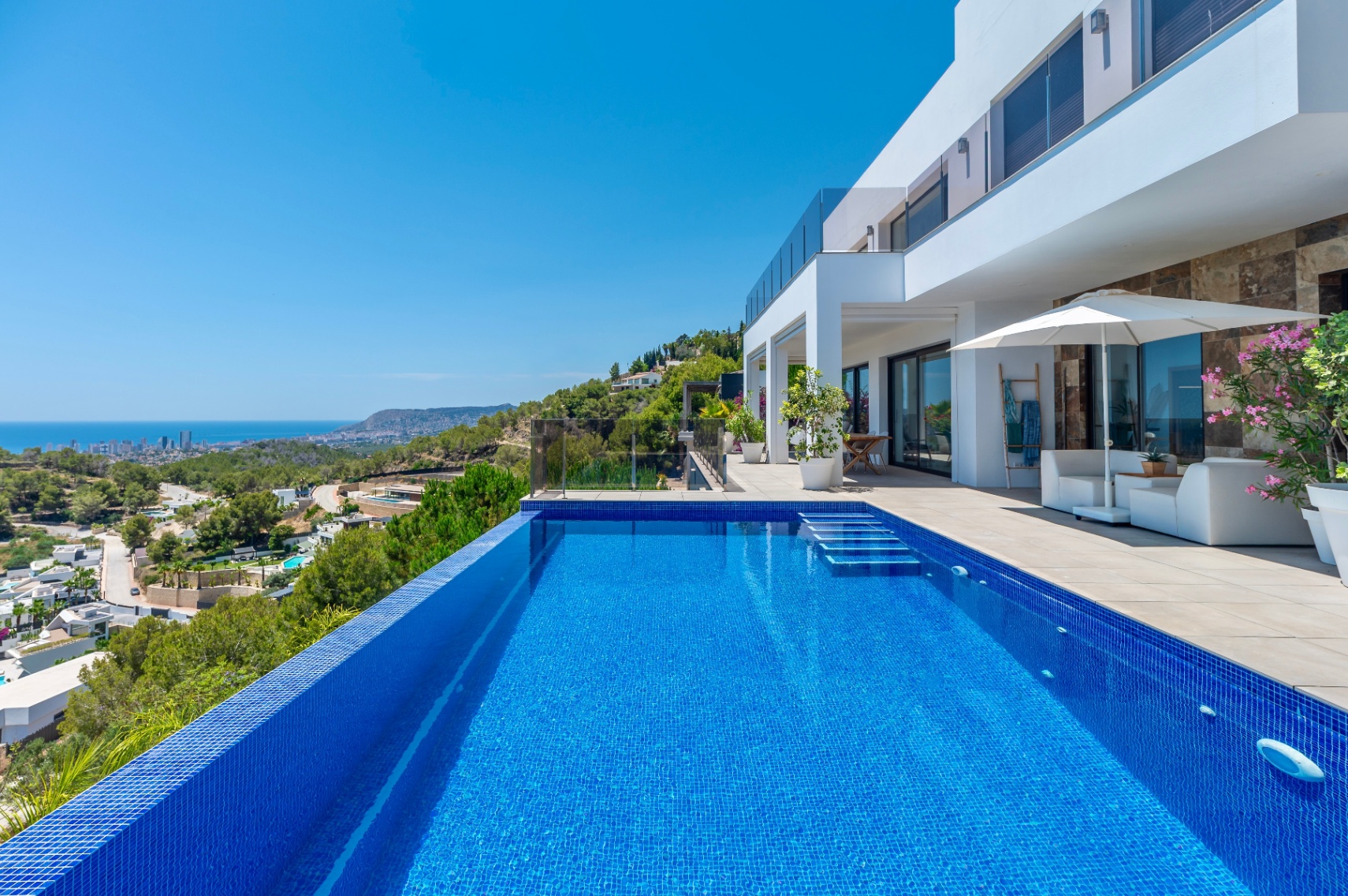 Infinity pool with blue mosaic tiles and integrated steps, bordered by a spacious terrace featuring white lounge furniture and panoramic coastal and city views.