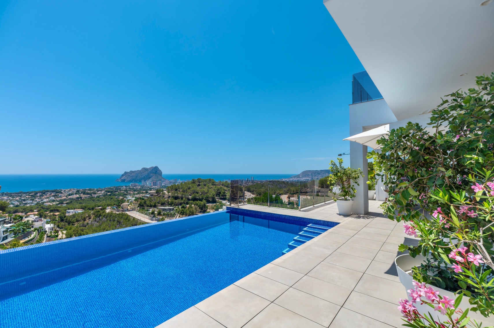 Infinity pool with vibrant blue mosaic tiles and an expansive light-tiled terrace. Features unobstructed panoramic sea views towards Peñon de Ifach, complemented by contemporary architecture and Mediterranean flora.