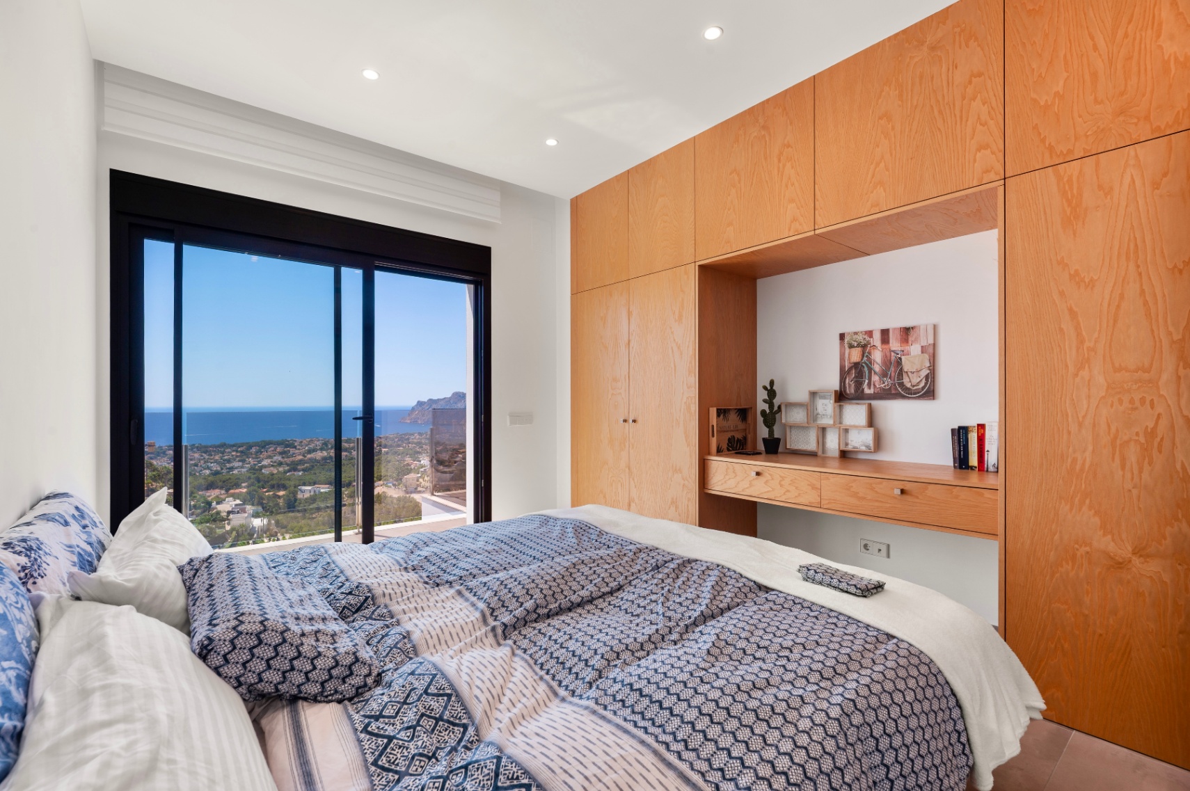 Master bedroom featuring expansive sliding glass doors offering panoramic sea and coastal views. Custom light wood cabinetry with an integrated desk provides elegant storage.