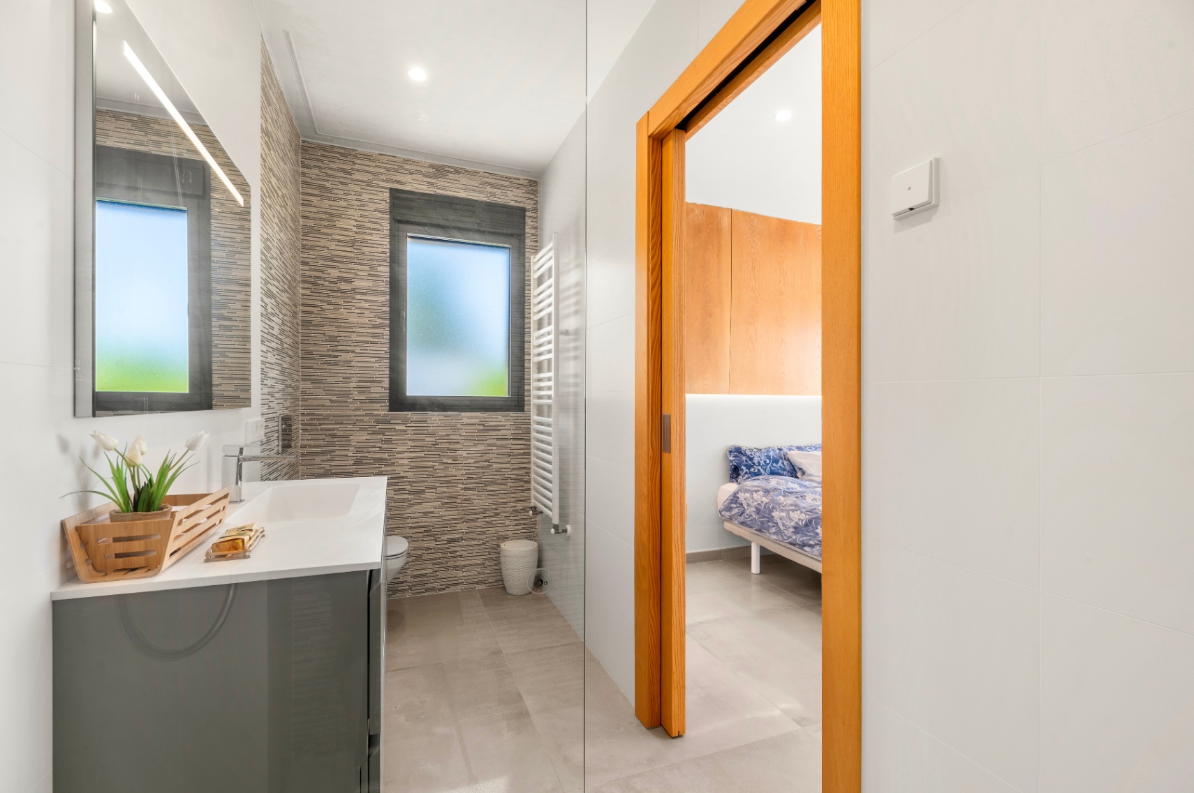 Modern bathroom featuring a grey vanity, illuminated mirror, and textured wall tiles. A frosted window and a white heated towel rail are visible, with direct access to a bedroom.