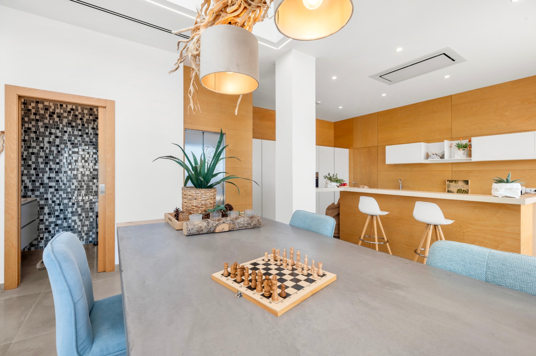 Modern dining area featuring a concrete-style table, light blue chairs, and a unique natural wood chandelier. An open kitchen with light wood paneling and a bar is visible, alongside a mosaic-tiled bathroom.