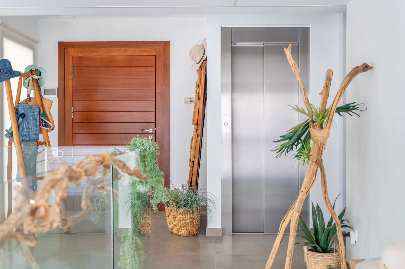 Modern interior landing featuring a sleek wooden door and a stainless steel elevator. The space is elegantly decorated with natural elements like driftwood, potted plants in woven baskets, and a glass railing.
