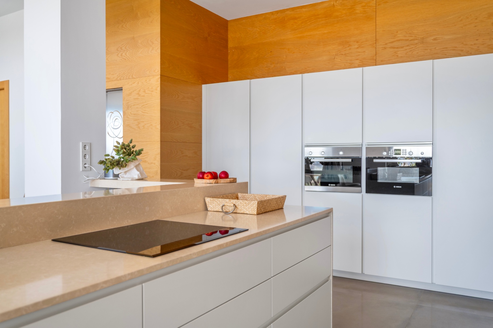 Modern kitchen featuring a sand-toned countertop with integrated induction hob. White handleless cabinets house two built-in Bosch ovens and a striking wooden accent wall.