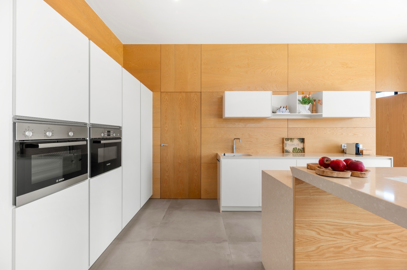 Modern kitchen featuring integrated white cabinetry with Bosch ovens. A central island with a light stone countertop and light wood paneling on walls and ceiling define the space.