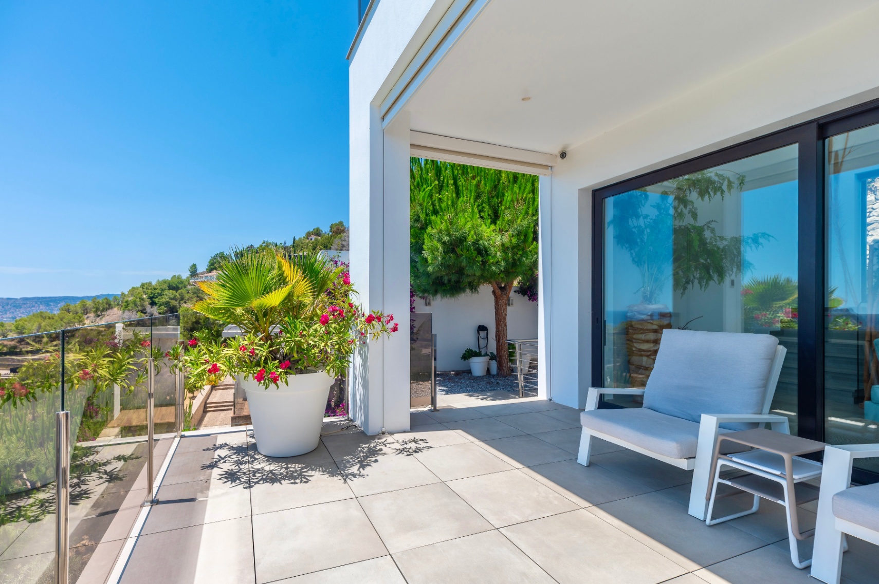 Modern outdoor terrace with light grey tiled flooring and a glass railing, offering a clear view of the verdant hillside. Features comfortable outdoor seating and vibrant potted plants under bright natural light.