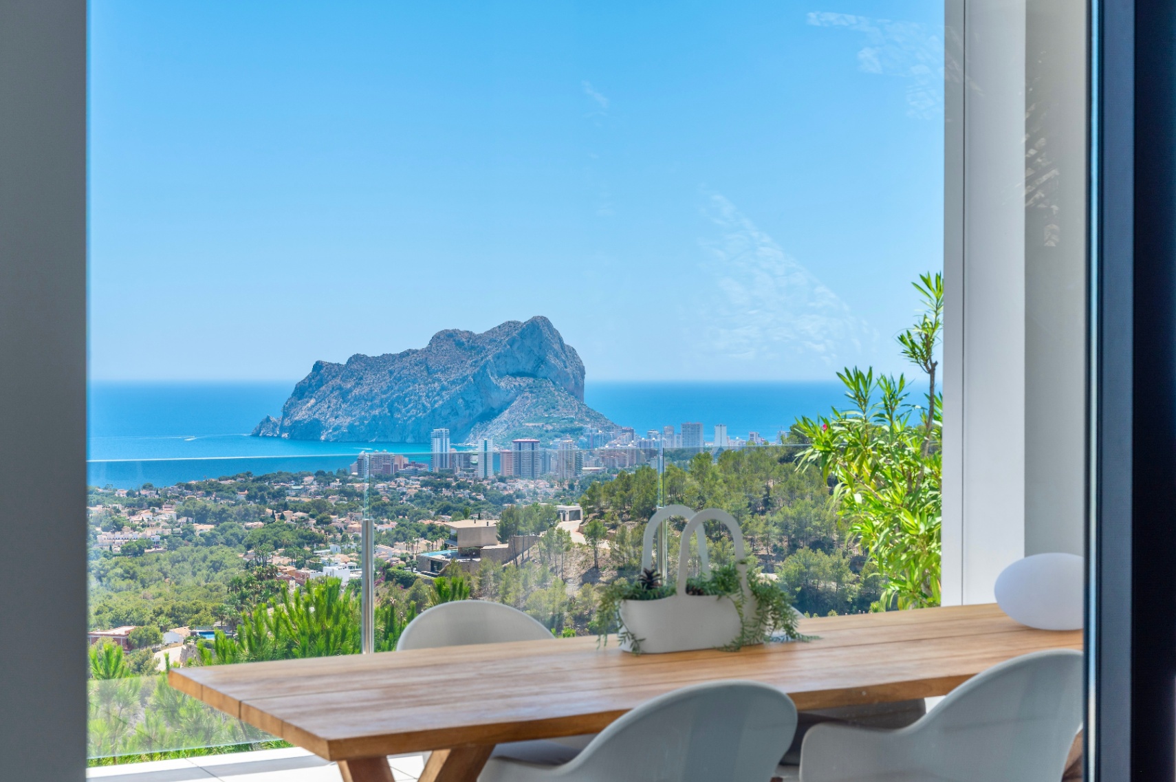Outdoor dining terrace featuring a wooden table and modern white chairs, offering panoramic views of Peñon de Ifach, the Mediterranean Sea, and Calpe's coastline.