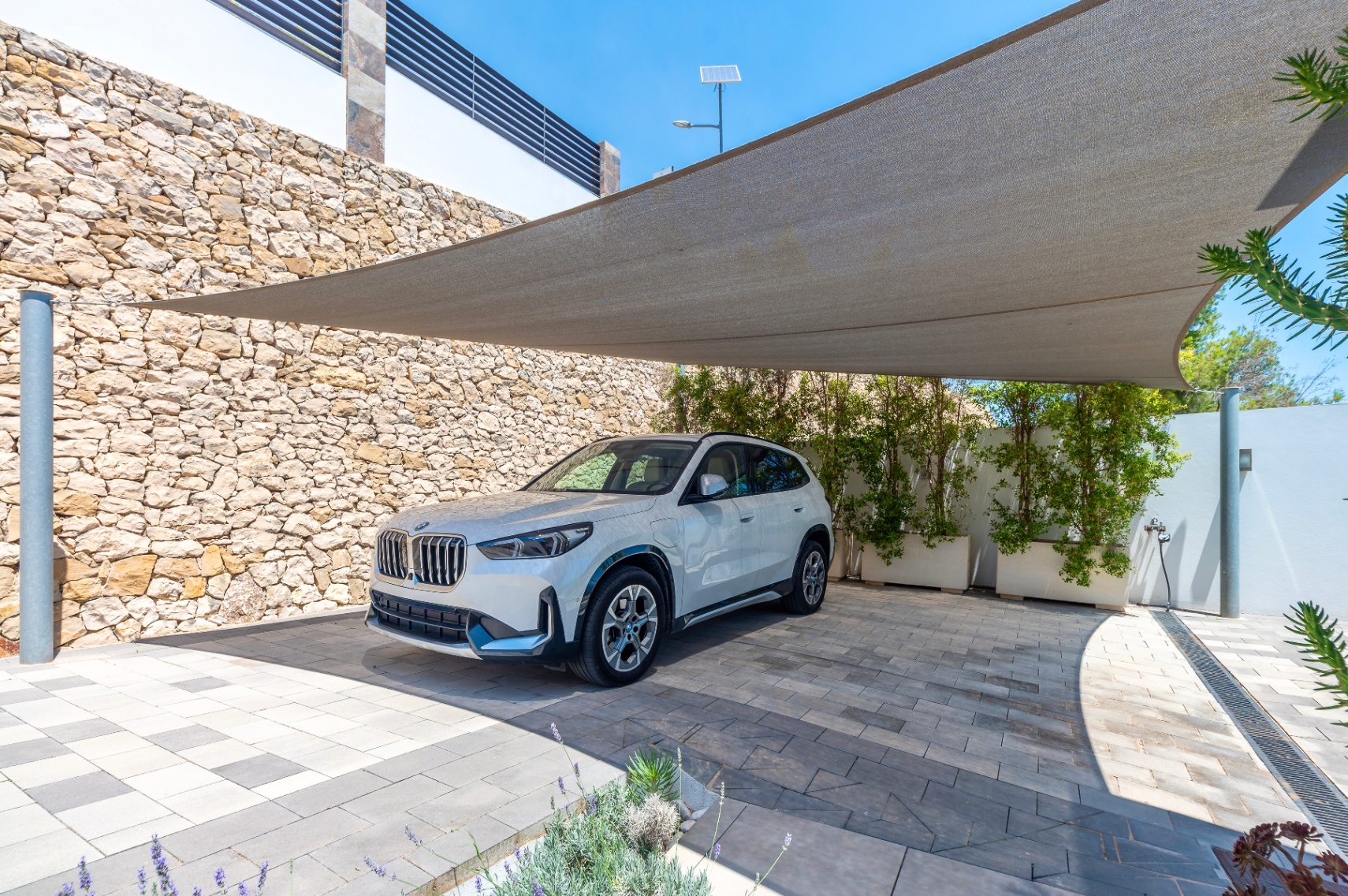 Outdoor parking area featuring a large shade sail over a patterned paved driveway. A natural stone wall and green planters define the space, with a white SUV parked.