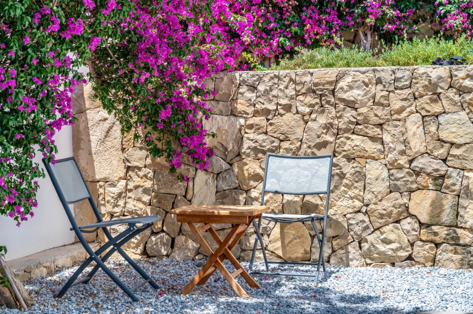 Outdoor seating area featuring two grey folding chairs and a wooden table on gravel. A natural stone wall is adorned with vibrant magenta bougainvillea.