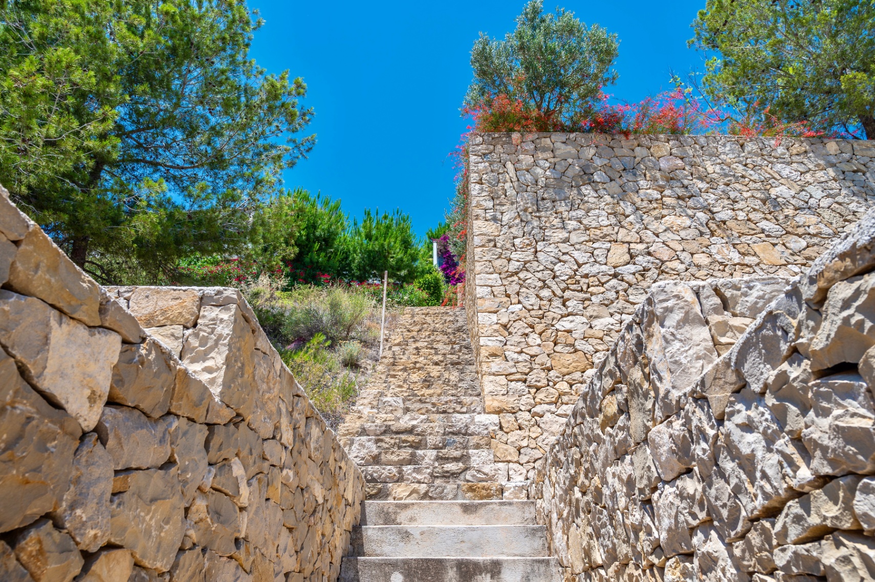Outdoor stone staircase ascending between dry-stacked natural stone walls, surrounded by lush Mediterranean trees and flowering shrubs under a clear blue sky.