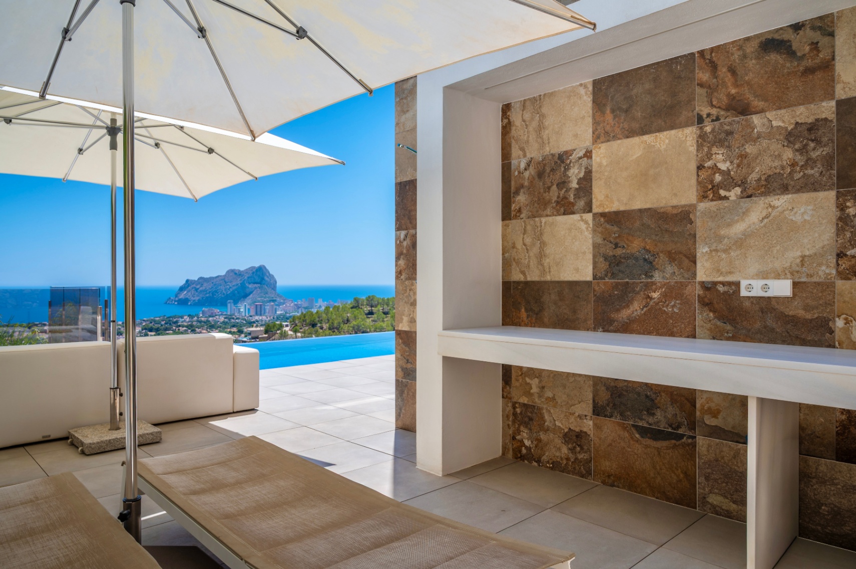 Outdoor terrace with a built-in counter featuring multi-toned natural stone-look tiles. Offers panoramic sea views, an infinity pool, and the iconic Peñon de Ifach.
