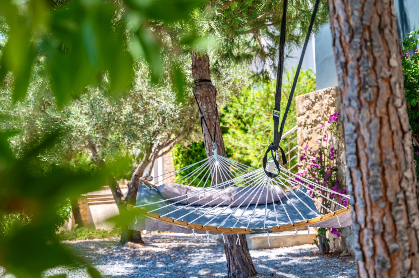 Padded grey hammock with a cushion, suspended between lush trees on a gravel ground. A stone wall with purple flowers adorns the garden background.