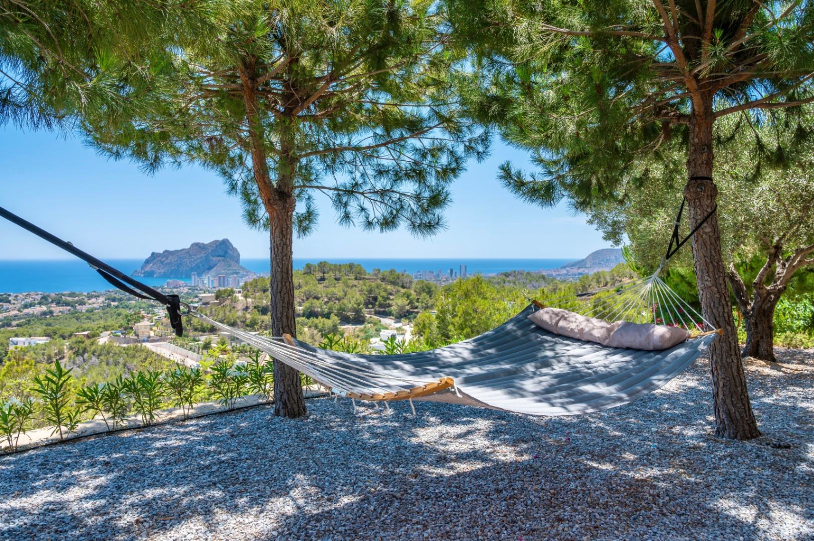 Padded hammock suspended between pine trees, providing a serene spot with panoramic views of the Mediterranean Sea and the prominent Peñon de Ifach. Gravel ground.