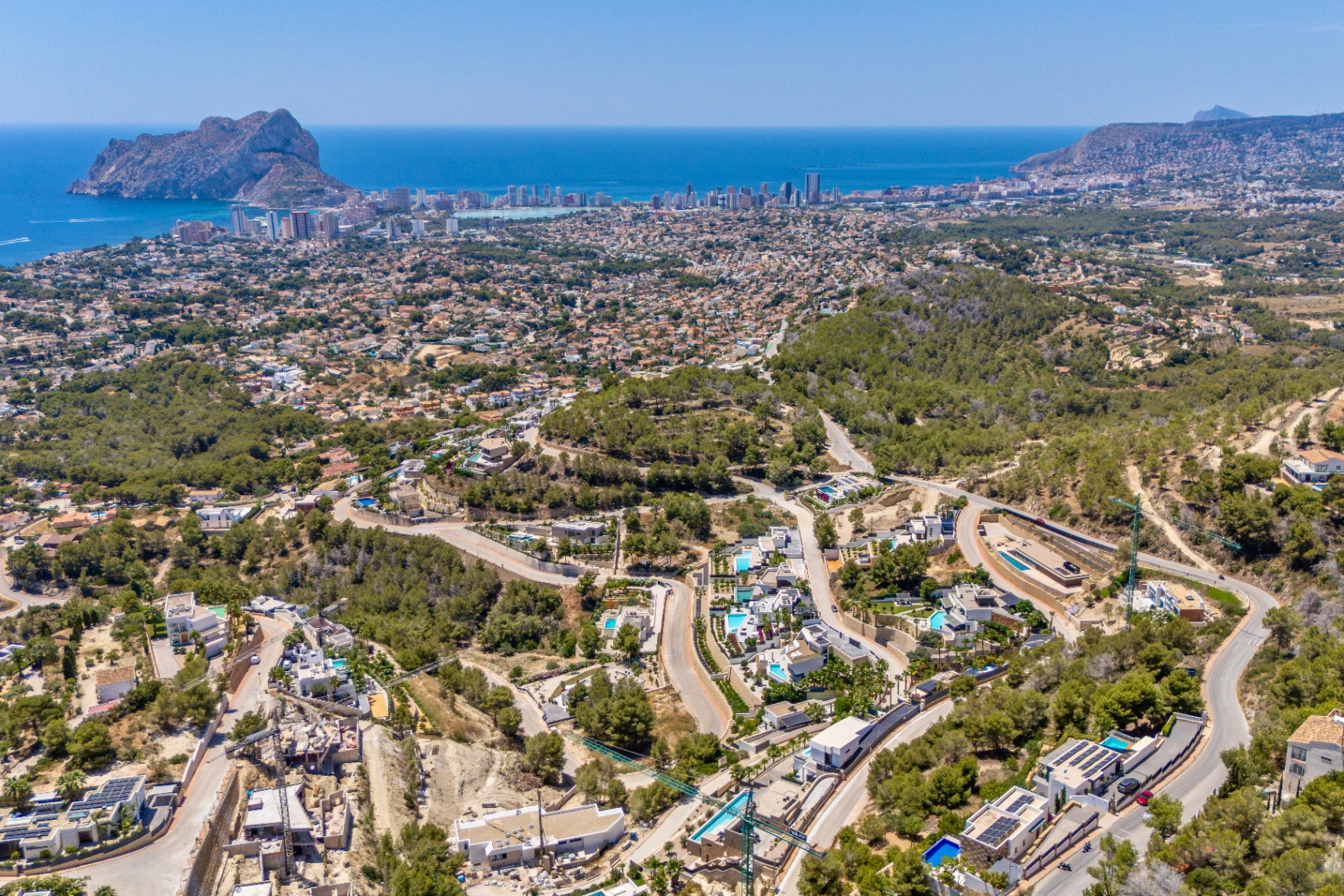 Panoramic aerial view of Calpe, showcasing the Peñon de Ifach, the coastline, and a residential area with villas and pools amidst green hills and winding roads.