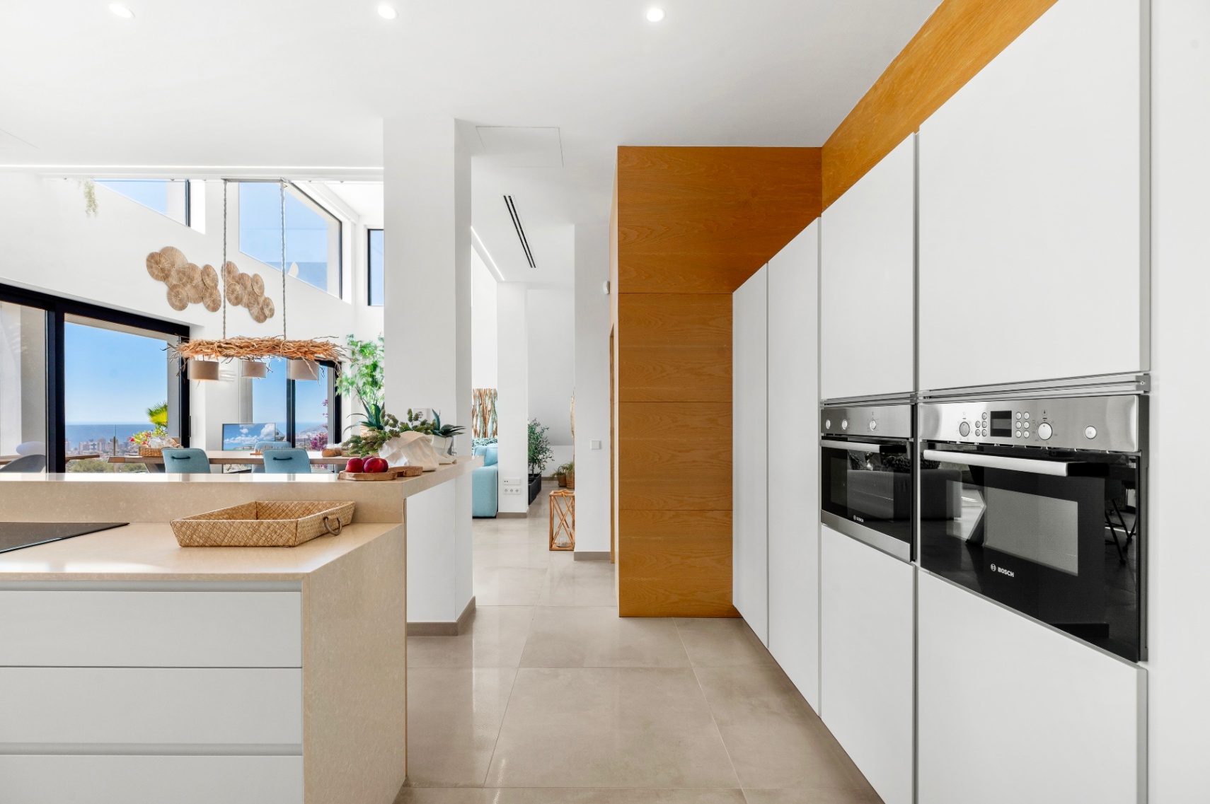 Sleek modern kitchen with integrated Bosch ovens, white handleless cabinetry, and a light stone island. The open layout extends to a dining area with panoramic sea views through large glass doors.