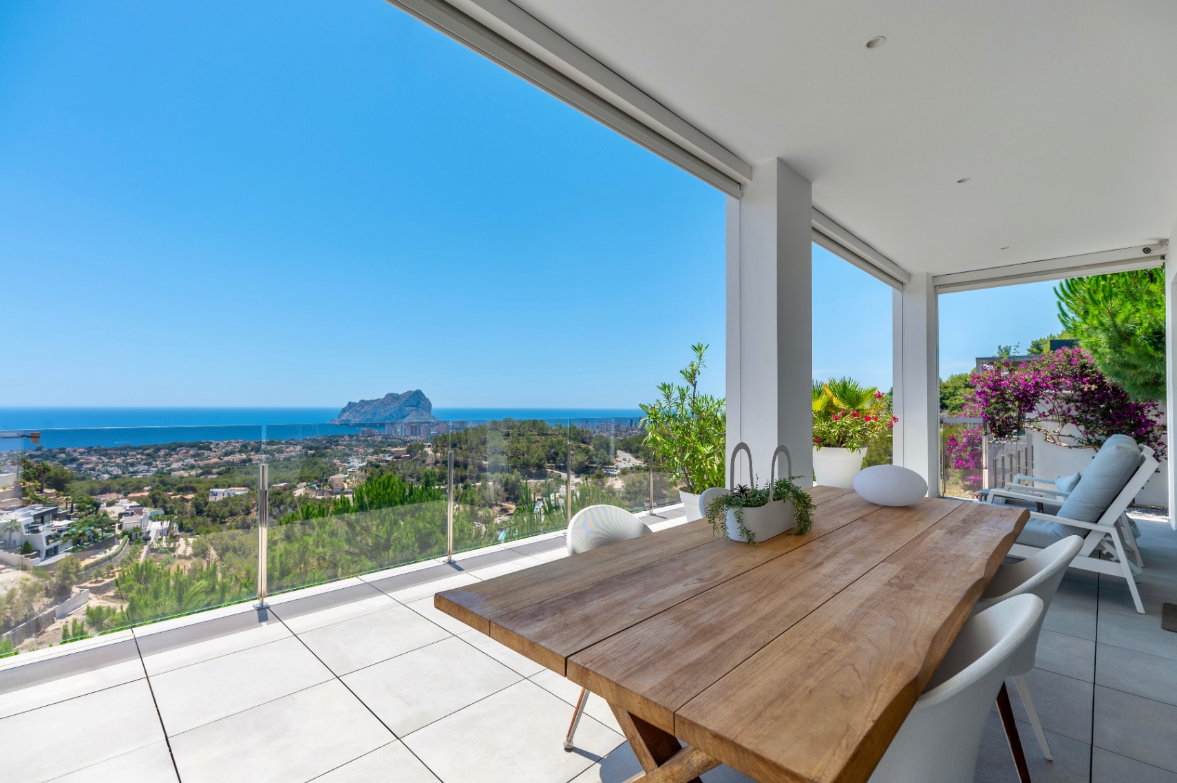 Spacious covered terrace featuring a solid wood dining table, white modern chairs, and light grey tiles. Offers panoramic sea views of Calpe and the iconic Peñon de Ifach through a clear glass railing.