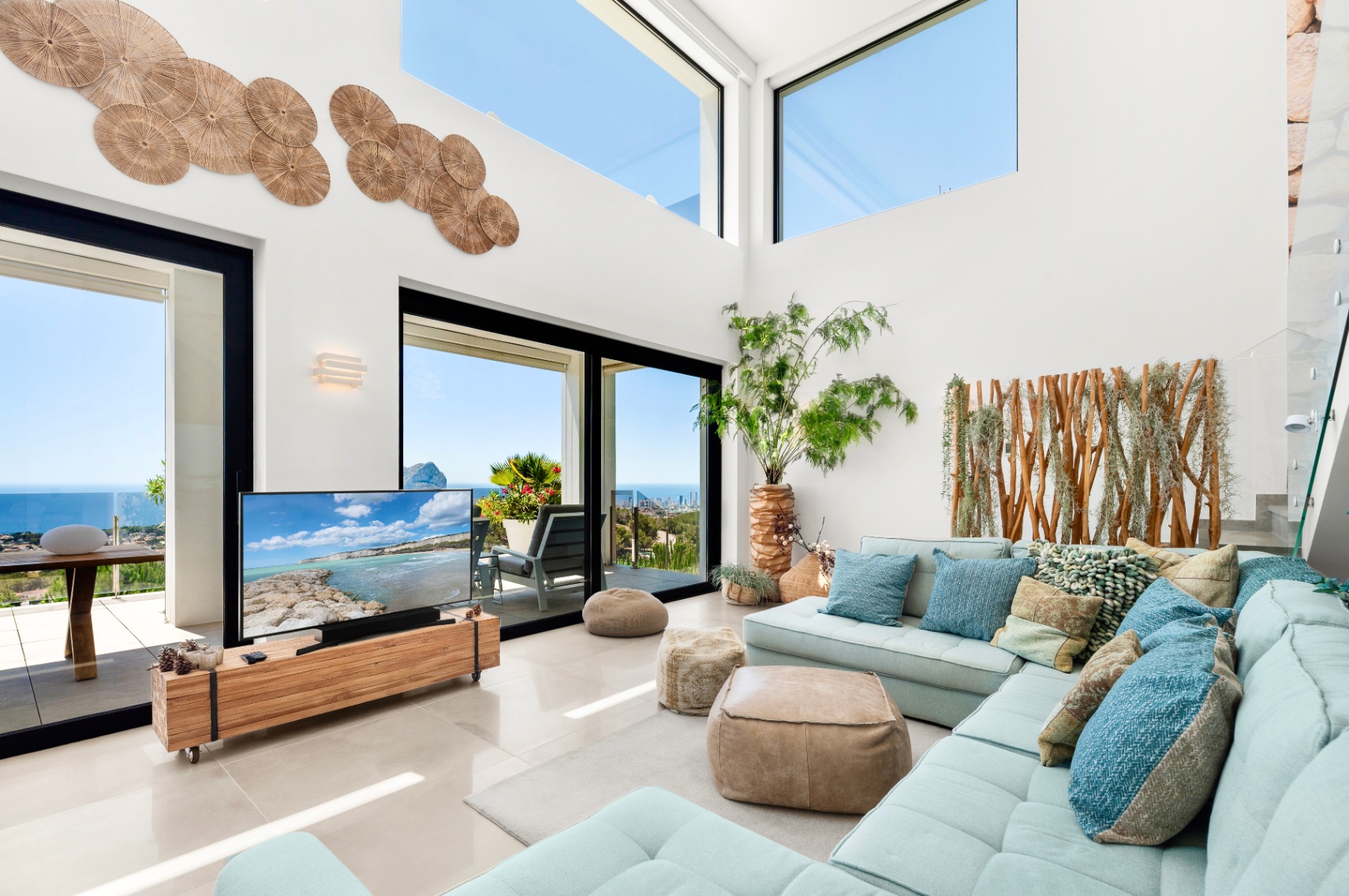 Spacious living room featuring high ceilings, large glass doors, and upper windows framing panoramic sea views. A light blue sectional sofa complements the natural wood accents and unique wall decor.