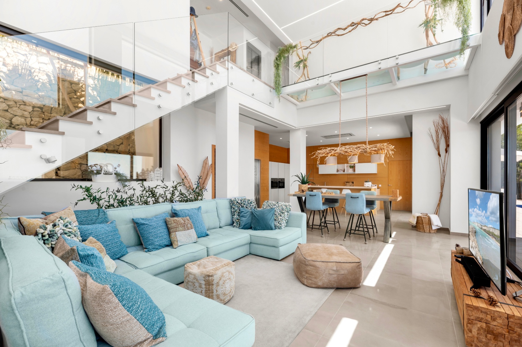 Spacious open-plan living area featuring a light blue sectional, a modern glass-railed staircase, and a kitchen with warm wood accents, all bathed in natural light.