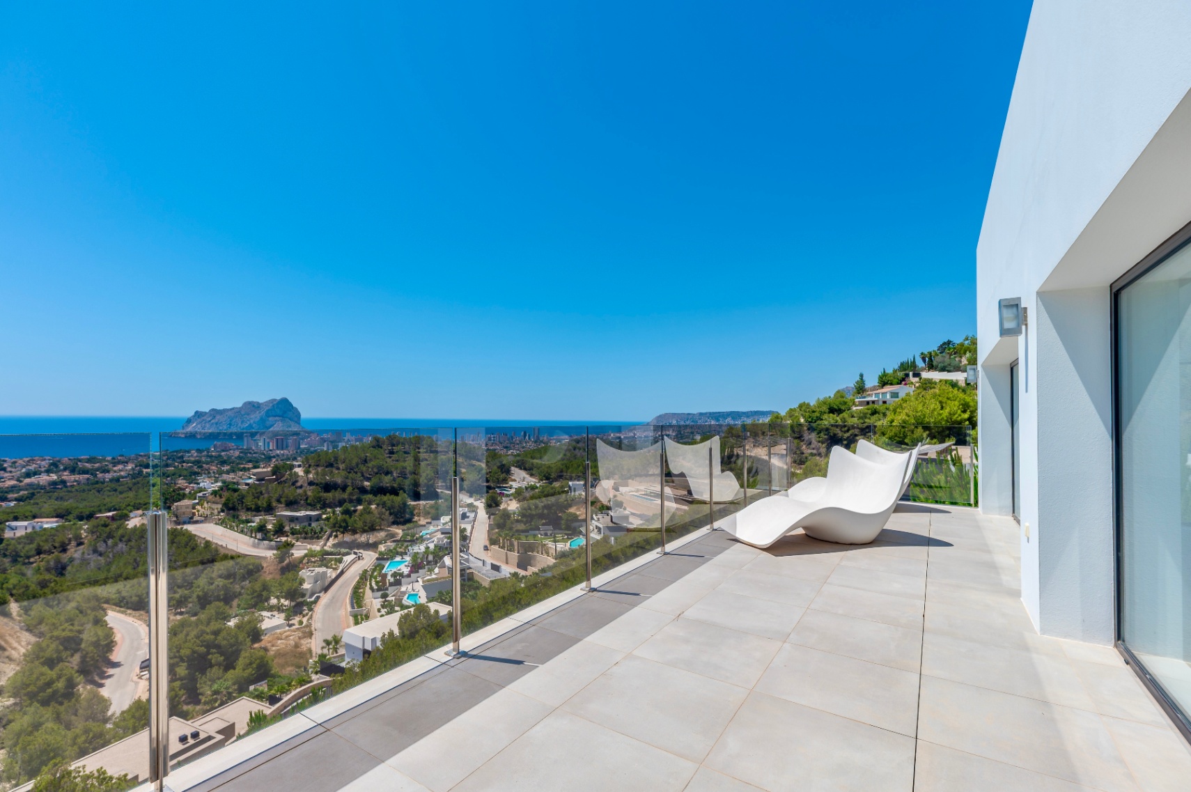 Spacious outdoor terrace featuring light-toned tiles, a modern white lounge chair, and a clear glass balustrade. Enjoy panoramic views of the Mediterranean Sea, the iconic Peñón de Ifach, and the Calpe coastline.