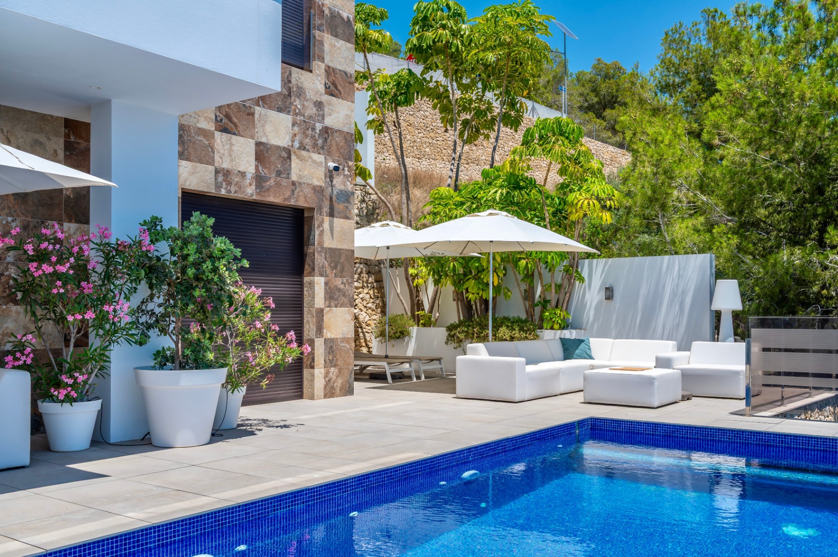 Spacious terrace featuring a blue mosaic pool and white outdoor furniture under umbrellas, complemented by a stone facade and lush Mediterranean greenery.