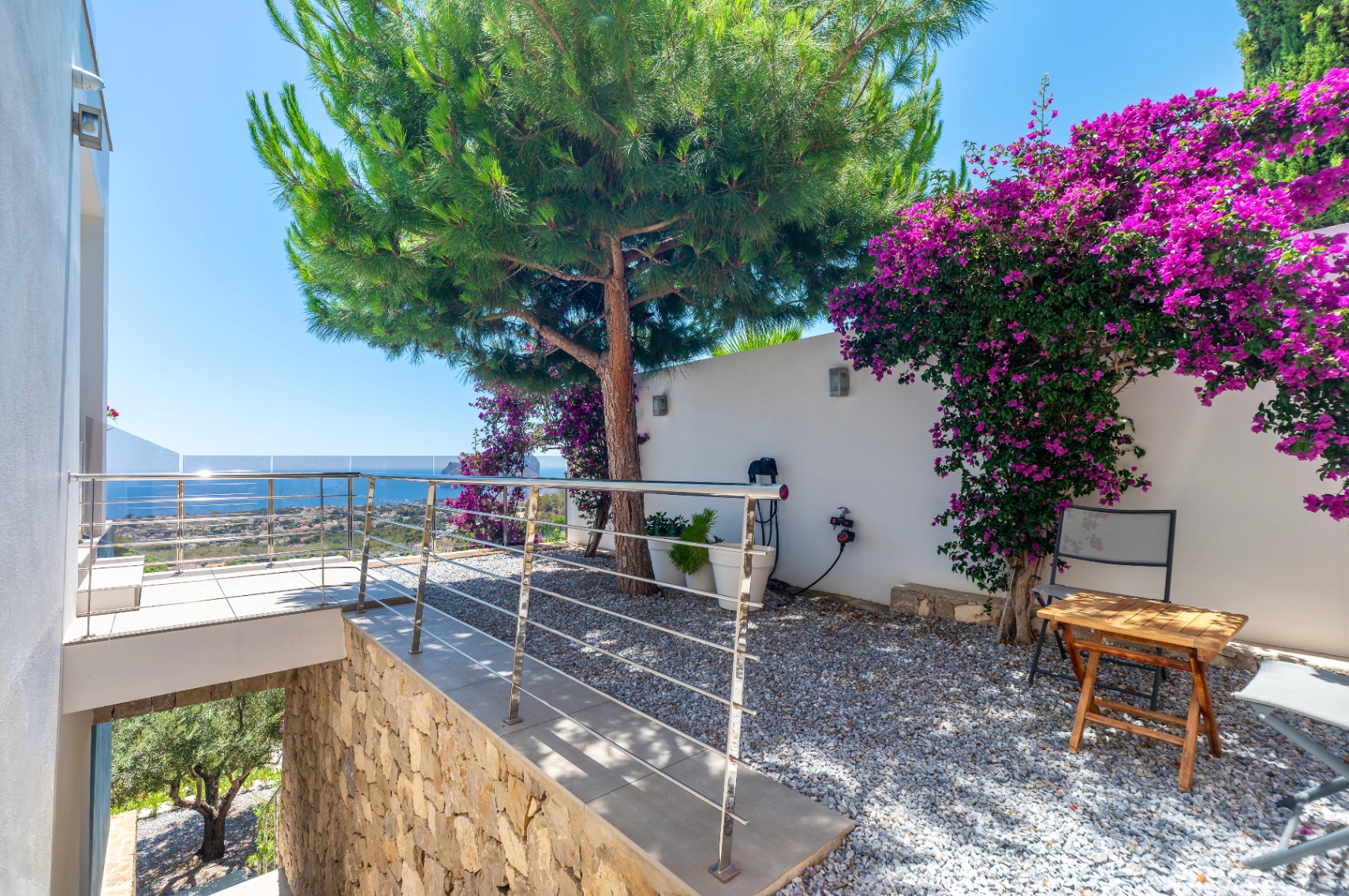 Terrace with modern stainless steel railing and glass, offering panoramic sea views. Adjacent gravel garden features a large pine tree and vibrant purple bougainvillea against a white wall.