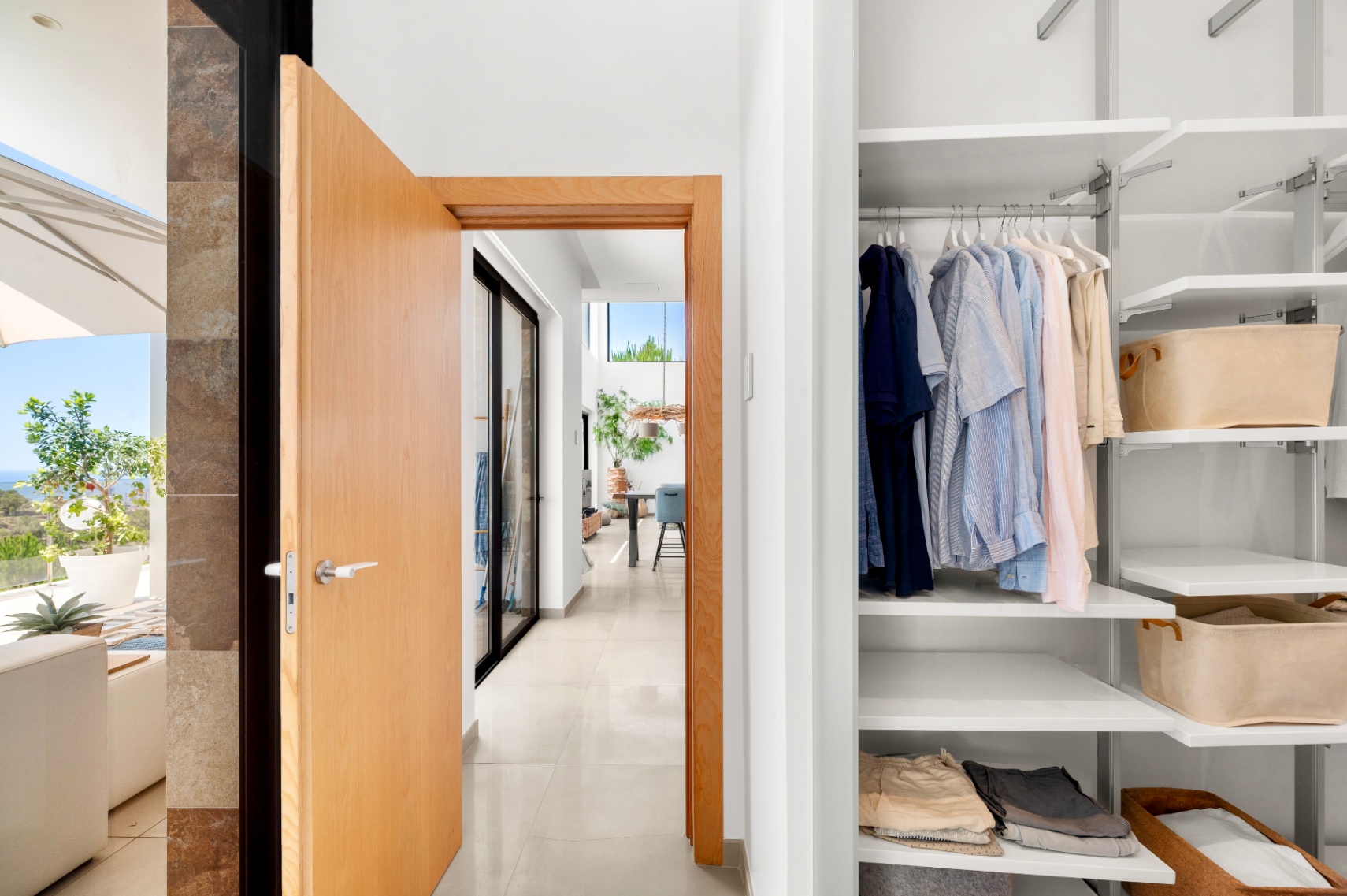 This photo captures a modern hallway entrance with an open light wood door, revealing a built-in white closet with adjustable shelves and a glimpse of a sunlit outdoor terrace with sea views.