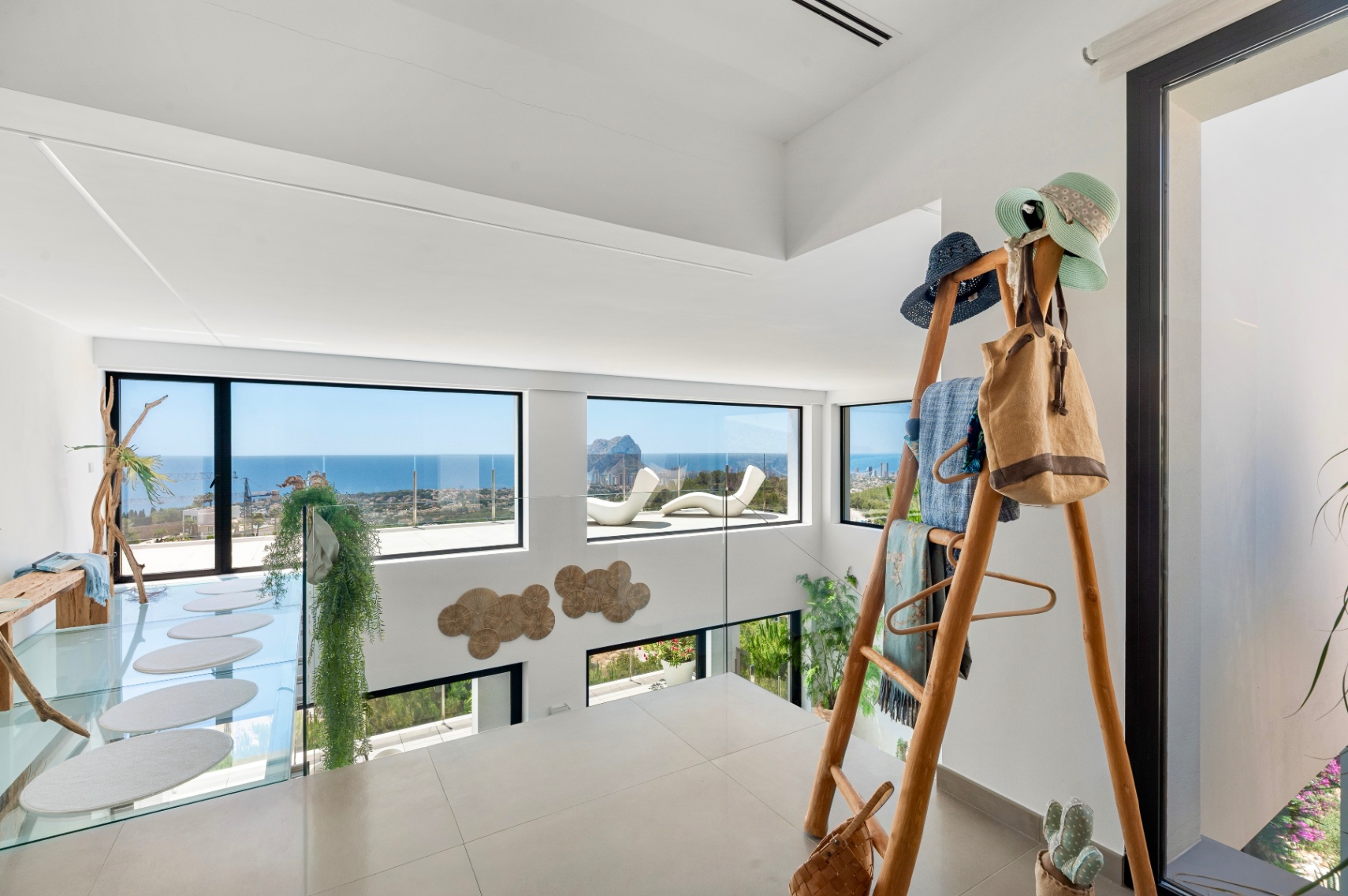 Upper level with light-toned tiled floor and glass balustrade. Expansive windows frame panoramic sea views, including Peñon de Ifach. Features a glass walkway with oval steps and a unique wooden rack.