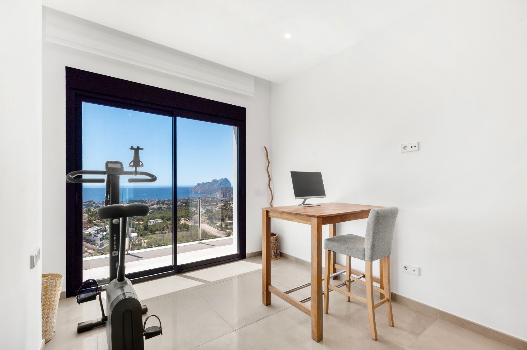 Versatile room featuring a home office setup with a wooden high desk and a fitness bike, both positioned to enjoy the expansive sea views and the iconic Peñon de Ifach through the large sliding glass doors.