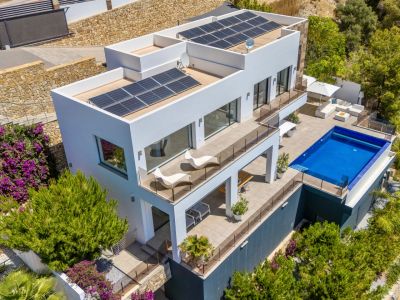 Aerial view of a modern white villa featuring multiple terraced levels, a rectangular swimming pool, and extensive outdoor lounge areas. Solar panels are visible on the flat roofs, set amidst lush Mediterranean landscaping.