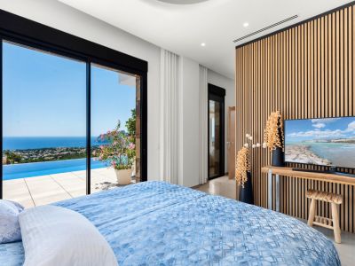 Bedroom featuring expansive sliding glass doors that open to an infinity pool and panoramic sea views. A modern slatted wood wall serves as a backdrop for the TV, complementing the light interior.