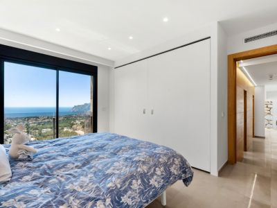 Bedroom with full-height sliding glass doors showcasing panoramic sea and town views. Features include integrated white wardrobes, recessed lighting, and a light tiled floor.