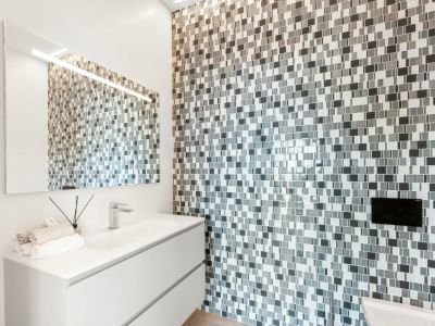 Contemporary bathroom featuring a white floating vanity with integrated sink and chrome faucet. The accent wall is clad in a grey and white mosaic tile, complemented by an illuminated mirror.